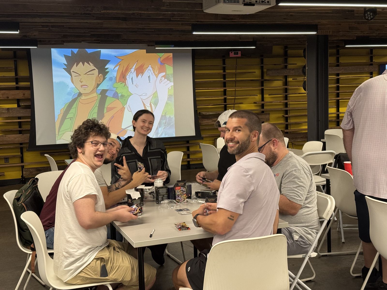 People smiling and opening packs at a community game night table at Tabbris, with Ash and Misty on the projector