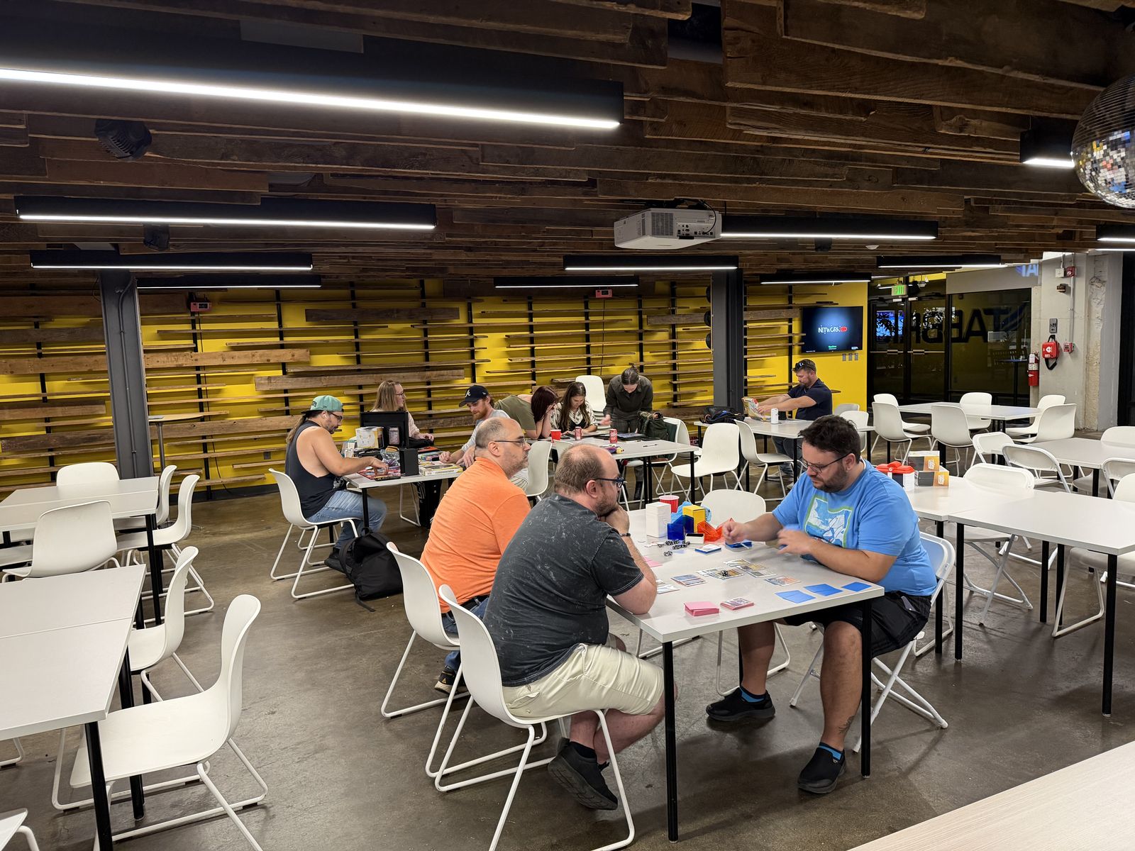 A play night in the Tabbris event space, with several tables of players and a projector at the front of the room