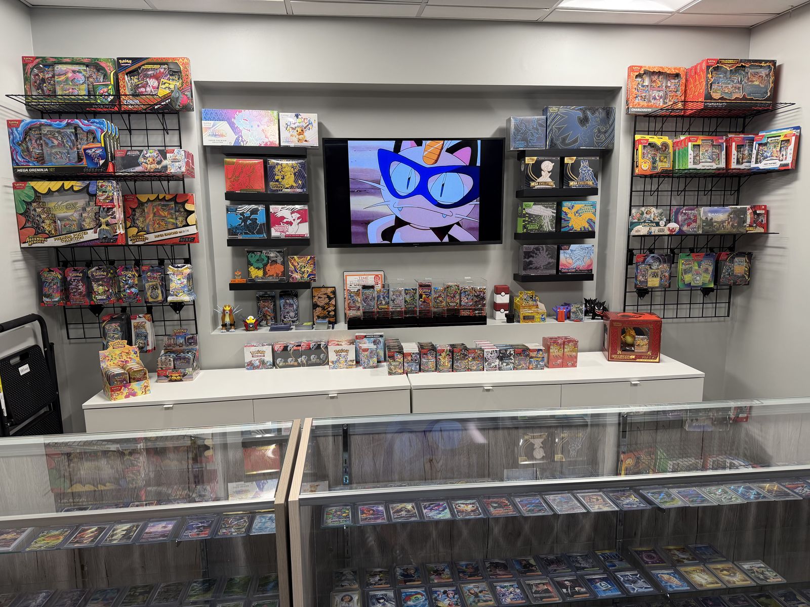 The shop’s main wall of sealed Pokémon tins, Elite Trainer Boxes, and booster bundles, with a TV and a glass case of singles in front