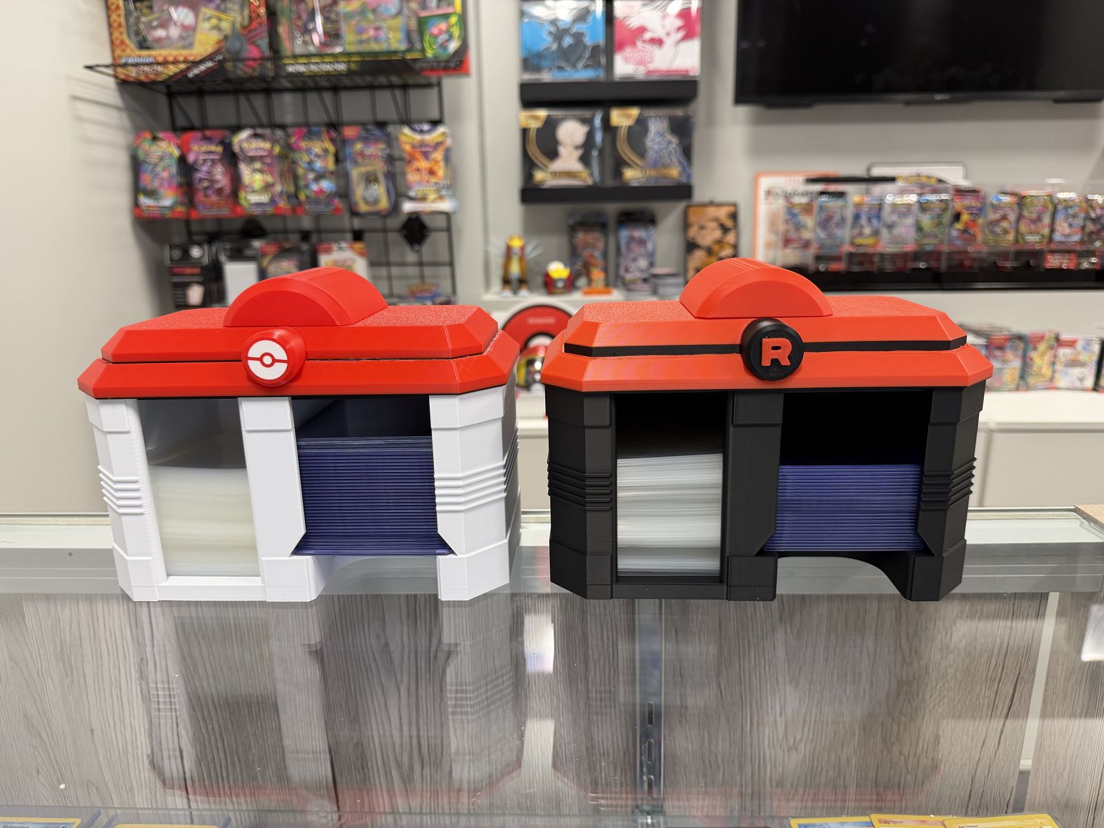 Two 3D-printed card-sleeve holders on the shop counter: a Poké Ball and a red-and-white Team Rocket design
